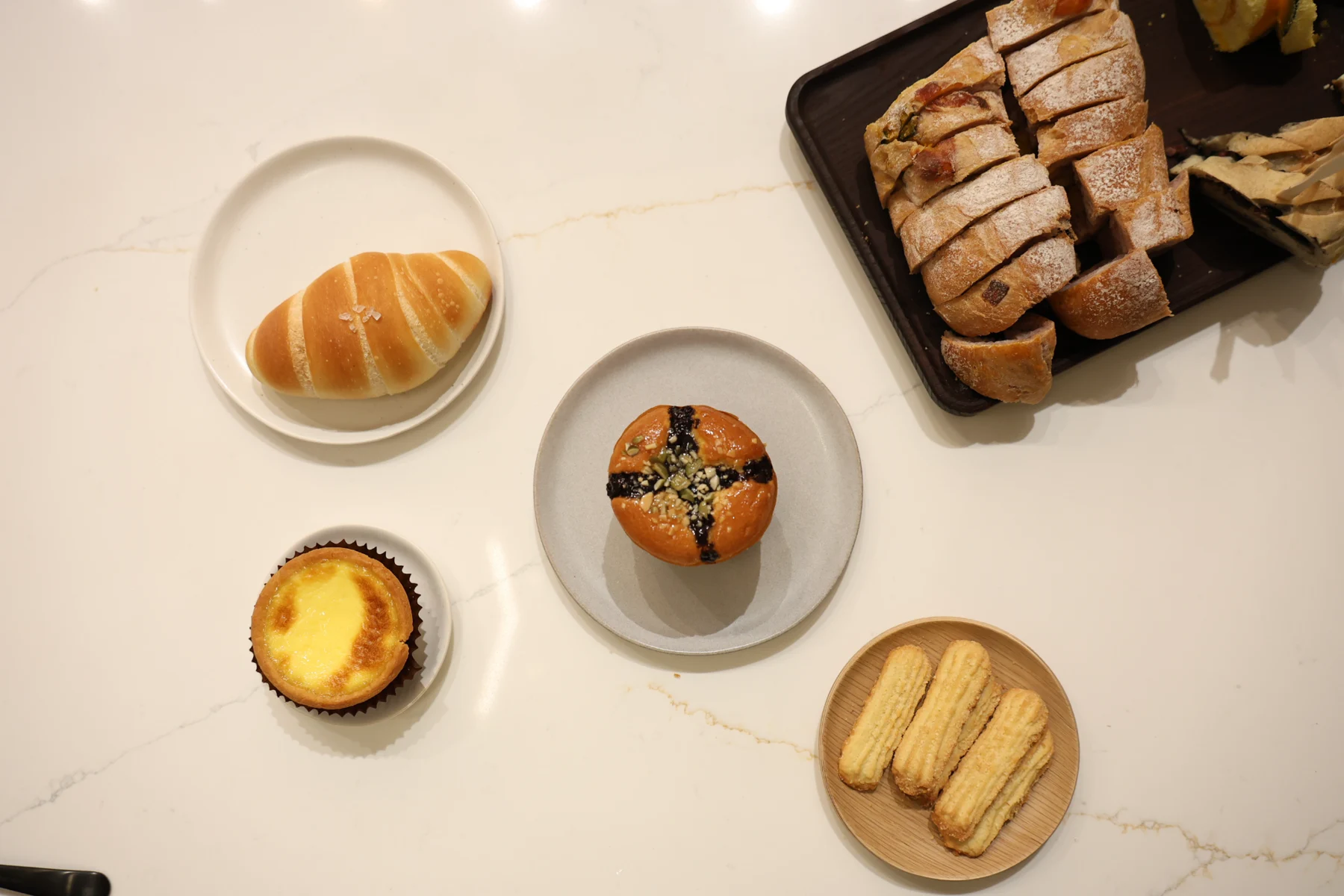 An overhead view of breads, an egg tart, and sweet treats arranged on a light table